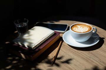 Latte coffee and reading book on wooden table with sunlight and diary for writing note, relax during a day after work in cafe