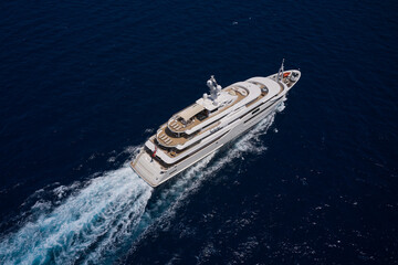 Aerial view of a white mega yacht with five floors and a wooden deck, moving fast on dark blue water. Yacht for millionaires in motion. Top view of a modern, luxurious yacht speeding on water.