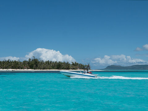 Speedboat cruising through the emerald tropical sea, with palm-lined beaches and sandy shores of Managaha Island in Saipan. Clear blue skies, scattered clouds, and a stunning island paradise scene