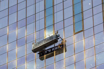 Male window cleaner cleaning glass windows on modern building high in the air on a lift platform. A worker washes windows in a building from a crane platform