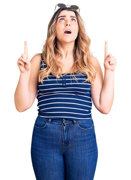 Young caucasian woman wearing casual clothes and sunglasses amazed and surprised looking up and pointing with fingers and raised arms.
