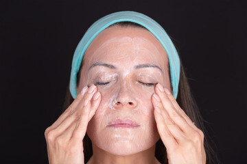 Close-up portrait of a woman performing a facial make-up removal routine, with closed eyes and a light blue headband. Black background. Conceit of facial cleansing, personal care and beauty routine