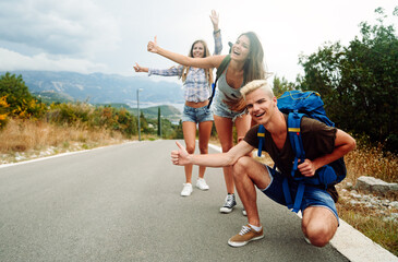 Cheerful interracial friends standing on road showing hitchhiking gestures. Tourism traveling