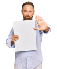 Handsome middle age man holding blank empty banner with open hand doing stop sign with serious and confident expression, defense gesture
