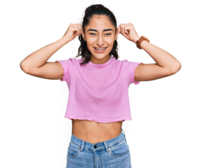 Hispanic teenager girl with dental braces wearing casual clothes smiling pulling ears with fingers, funny gesture. audition problem