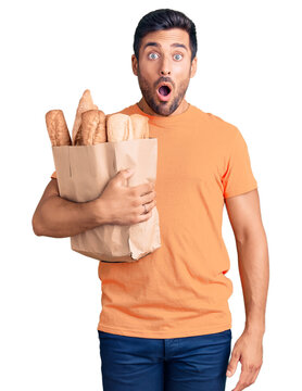 Young hispanic man holding paper bag with bread scared and amazed with open mouth for surprise, disbelief face
