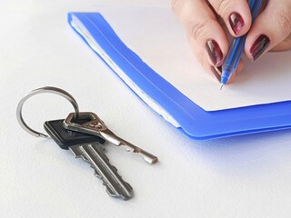 A woman writes with a pen on a piece of paper, keys are lying on the table next to her. Housing issuance to low-income families. Real estate rental. Purchase and sale of property.