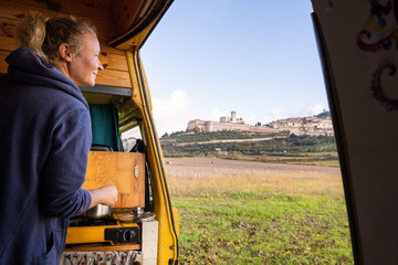 Young woman cooking breakfast in vintage camper van near Assisi hilltop town in Umbria, Italy
