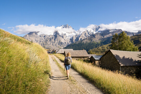 Woman hiking in Ecrins national park overlooking snowcapped Alps mountains with ancient stone houses along the path.