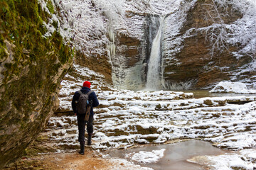 a male tourist with a backpack travels along a forest stream with waterfalls on a winter day