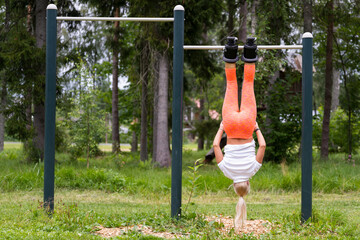 A young athletic girl in gravity boots hangs on a horizontal bar outdoors on a summer morning. Back view.