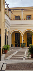 Obraz premium Yellow courtyard with arched colonnades, potted plants, and geometric tiled flooring; a second-floor balcony features potted flowers and simple railings;