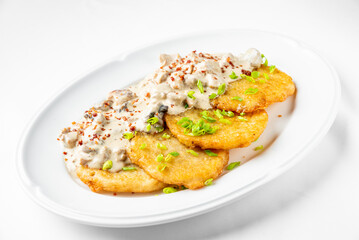 A plate of crispy golden potato pancakes topped with a creamy mushroom sauce and garnished with chopped green onions, showcasing delicious comfort food on a white background.