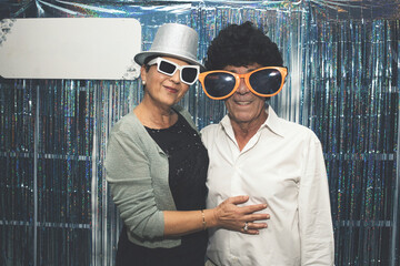 Senior couple wearing oversized, humorous glasses and a playful hat, posing joyfully for a photo booth picture against a shiny silver curtain backdrop, capturing festive memories together