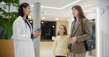 Obraz premium Mother and Daughter Consulting an Asian Female Doctor in a Modern Hospital Reception Area. They are Standing in a Bright Interior with Indoor Plants, Circular Seating and Elevators