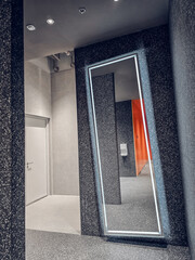 Featuring a large,angled full-length mirror framed in silver or metallic trim, leaning against a dark textured wall.The reflection in the mirror.The overall design gives a contemporary and stylish.