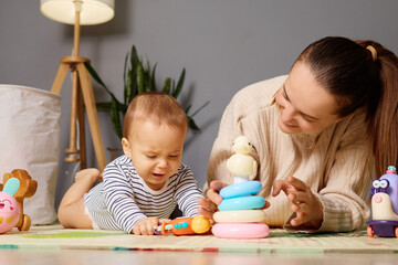 Obraz premium A mother sits beside her baby, guiding him as he plays with colorful stacking rings and toys on the floor. Warm light fills the cozy living room, creating a nurturing environment