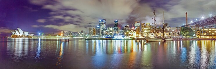 Nighttime panorama of Sydney Harbour showcasing the illuminated Sydney Opera House, city skyline, historic ships, and Harbour Bridge reflections on the water