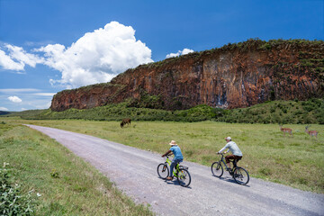Two cyclists ride along a dirt road in Hells Gate NP with a towering rocky cliff and wildlife in...