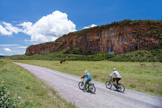 Two cyclists ride along a dirt road in Hells Gate NP with a towering rocky cliff and wildlife in the background