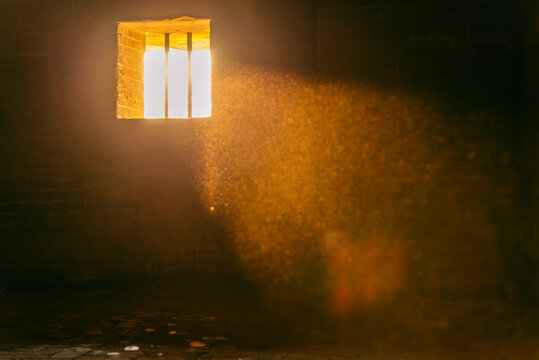 Golden rays of sunlight pierce through a barred window, casting a warm glow and revealing floating dust in a dim, abandoned space.