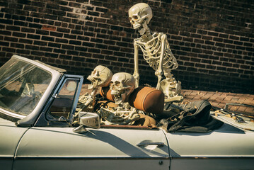 Skeleton crew decorates a vintage convertible for Halloween, parked against a brick wall backdrop.