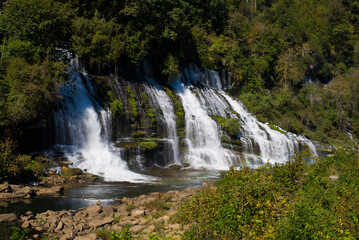 Fototapeta premium A stunning multi-tiered waterfall cascading down rocky cliffs surrounded by lush green foliage, captured on a sunny day.