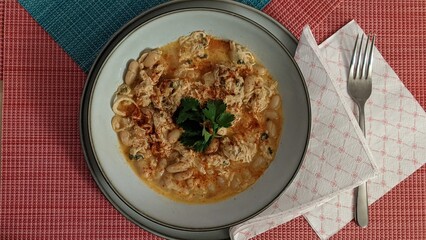 Top-down view of a bowl of white chicken chili with beans, garnished with fresh cilantro and paprika, served on a rustic red and blue tablecloth with a fork and napkin.
