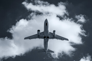 A commercial airplane flying overhead against a dramatic cloudy sky, captured in black and white.