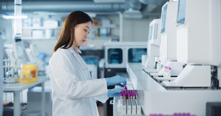 Naklejka premium Young Asian Scientist Carefully Prepares Test Vials for Genetic Analysis as Her Male and Female Colleagues Prepare Medical Equipment in the Background