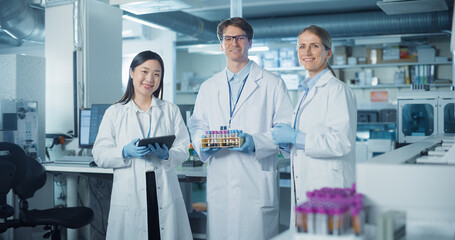 Portrait of a Diverse Research Team Standing in a Modern Laboratory in White Coats, Looking at Camera and Smiling. Female and Male Scientists Hold Test Samples and Tablet Computer