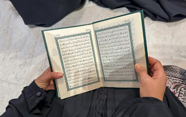 Arab woman pilgrim sits in Masjid al-Haram (the Sacred Mosque) and reads the Quran. A madinah manuscript. Close-up of an open book of the Quran in the hands of a girl in a black dress.