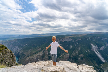 Obraz premium A young boy with blond hair stretches his arms wide while standing on a rocky cliff edge, enjoying the breathtaking view of the valley below