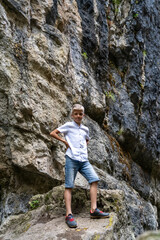 Young boy stands with hands on hips on a rock, surrounded by steep, textured cliff walls, enjoying a sunny day outdoors while exploring