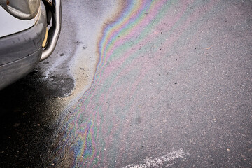 Obraz premium Rainbow gasoline or oil spill on wet asphalt road creating a colorful and polluting pattern near a parked car