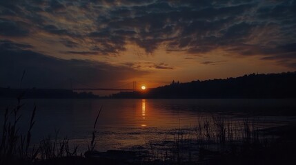Obraz premium A serene sunset over a river, with clouds reflecting vibrant colors and a bridge in the distance.