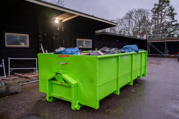 Bright green dumpster filled with construction debris near a black wooden building.