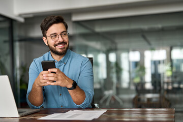 Busy happy young Latin business man holding smartphone sitting in office. Professional hispanic...