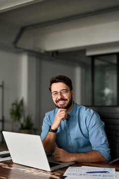 Vertical portrait of smiling young business man startup founder, happy Latin entrepreneur, confident business owner looking at camera using laptop computer technology working at corporate office desk.