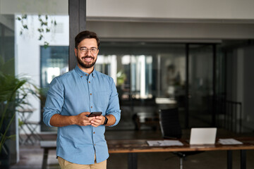 Obraz premium Happy professional Latin young business man looking at camera at work, portrait. Confident smiling businessman leader, male executive manager entrepreneur standing in office with smartphone in hands.