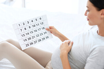Baby expecting concept. Young pregnant woman with big belly sitting on bed, holding calendar and...