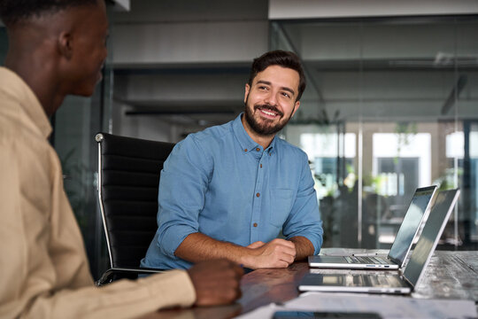 Two busy happy business men working on computers discussing online project data at work. Diverse professional workers developers team talking on finance software technology using laptops in office.