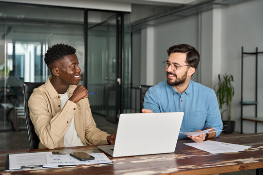 Two happy busy business men working on computers discussing online project at work. Diverse professional workers team talking on finance software technology analysis using laptops in office.