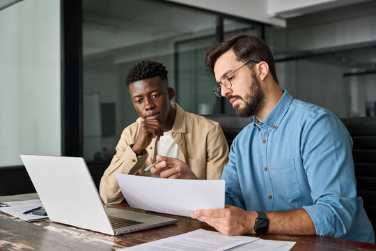 Two busy diverse male employees checking documents talking in office. Professional young business man manager financial advisor consulting client having conversation sitting at work meeting.