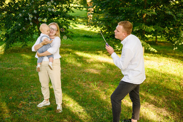 Cute young family standing in park, having fun together, mom holding baby in her arms, dad blowing...