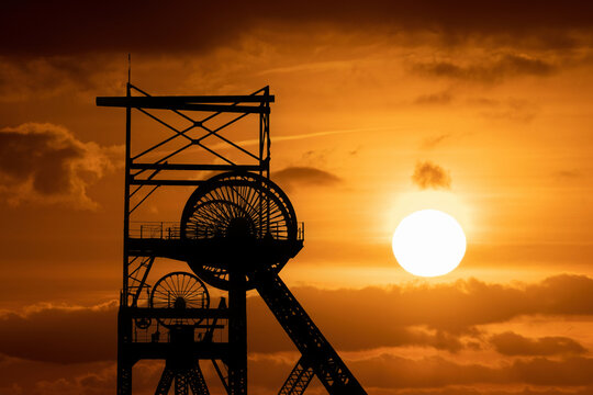 Silhouette of a historic coal mining headstock against a dramatic sunset sky with warm orange light highlighting Britain&rsquo;s industrial heritage and mining history.
