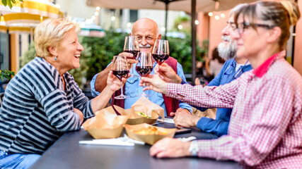 Happy senior friends having fun toasting and drinking red wine at dinner party - Retired people eating at restaurant patio together - Dining lifestyle concept with pensioners having fun together