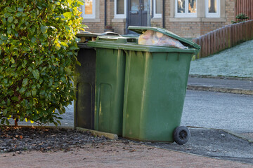 Overfilled Green Wheelie Bin with Lid Open on Paved Driveway
