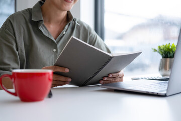Businesswoman sitting at desk on couch in workplace or at home working on laptop and analyzing data on charts and graphs and writing on papers to make business plan and strategies for company, 