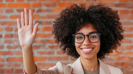 Smiling woman with curly hair waving in professional setting, showcasing confidence and friendliness. warm brick background adds welcoming touch